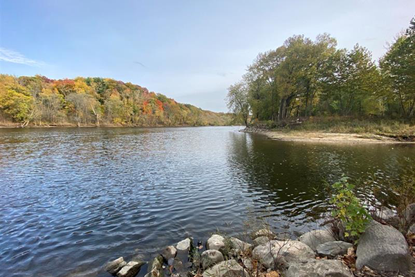 A large body of water stretches into the distance. It is surrounded by rocks, sandy banks, and trees that are just starting to show their autumn colors.