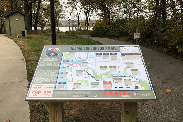 The Idema Explorer's Trail map stands next to a sidewalk that leads to a small outhouse and the riverside. The map shows how the trail leads through Connor Bayou.