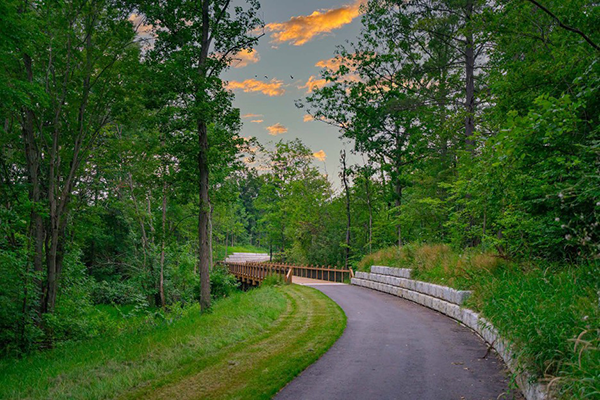 It is a calm summer sunset. A sidewalk leads away from the viewer toward a small footbridge and wooded area.
