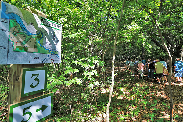 A group of people walk down a wooded path away from the viewer; A Stearns Creek Park map and trail marker stands in the foreground.