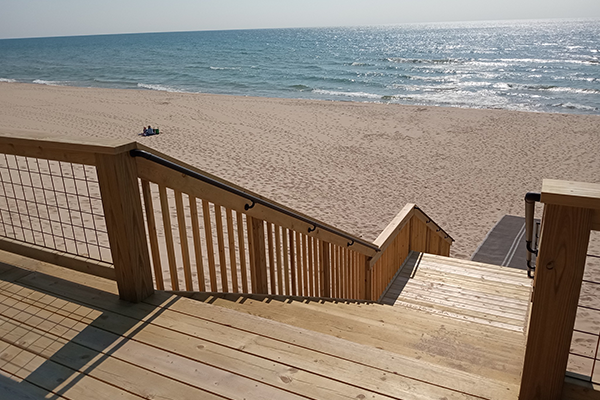 A wooden boardwalk leads down a hill to a beach, where two people sit on a blanket in the sand